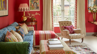  Bright and colourful room with red walls and a green and blue sofa. It has a cosy and comfortable style with bold lamps and a maximalist style.