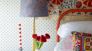  Close up of a red bed frame with brightly coloured cushions and lamp. A red and white striped candle sits on the bedside table.